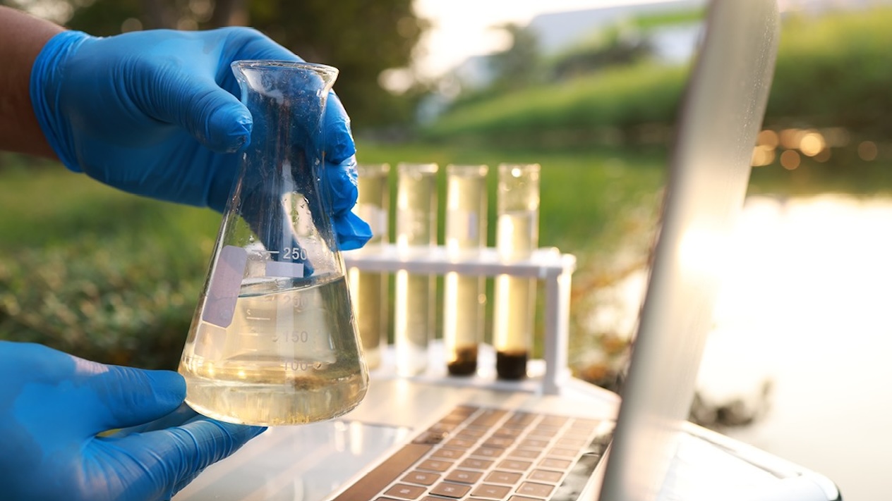 Gloved hands holding a water sample with a laptop, test tubes, and body of water in the background.