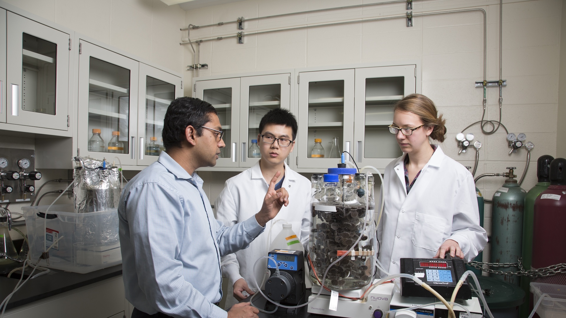 A professor and two lab coat-wearing researchers standing at a table-top machine