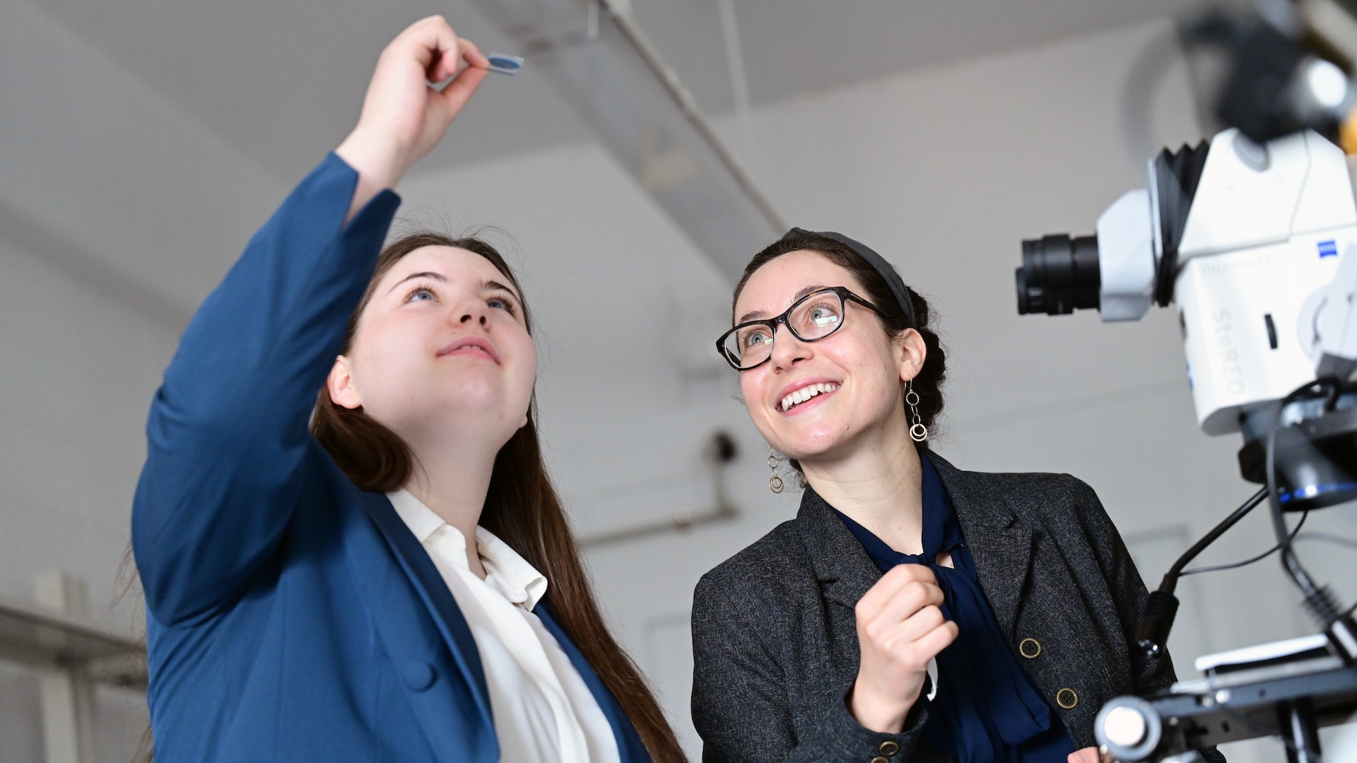 Two women. One woman holds and a microscope sample over their heads.
