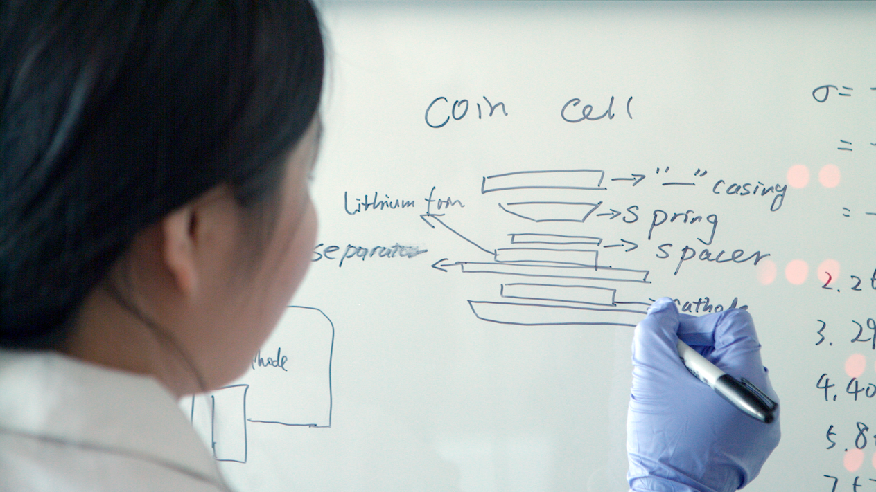 A student drawing a diagram that reads "coin cell" on a white board