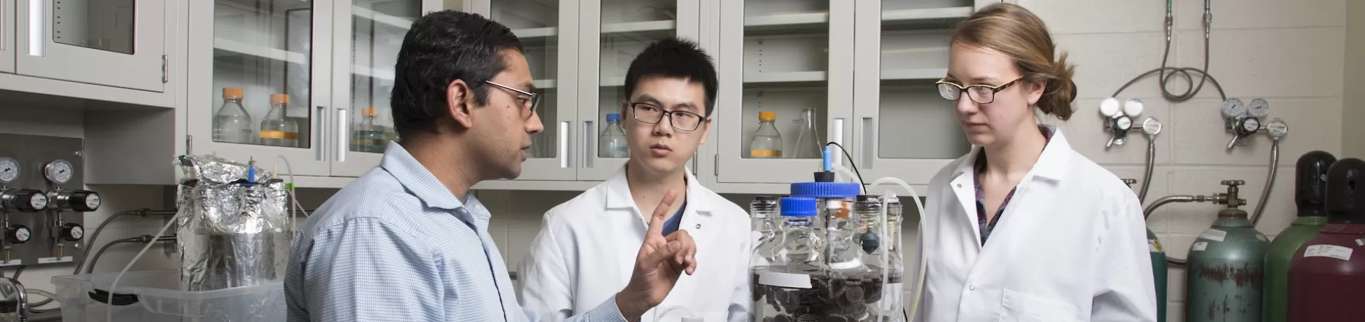 A professor and two lab coat-wearing researchers standing at a table-top machine