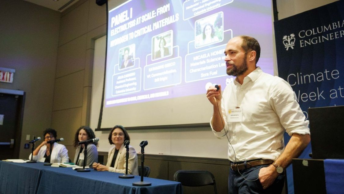 Dan Esposito speaking into a small wired microphone while standing beside three seated panelists, a projected presentation, and a Climate Week at Columbia Engineering banner.